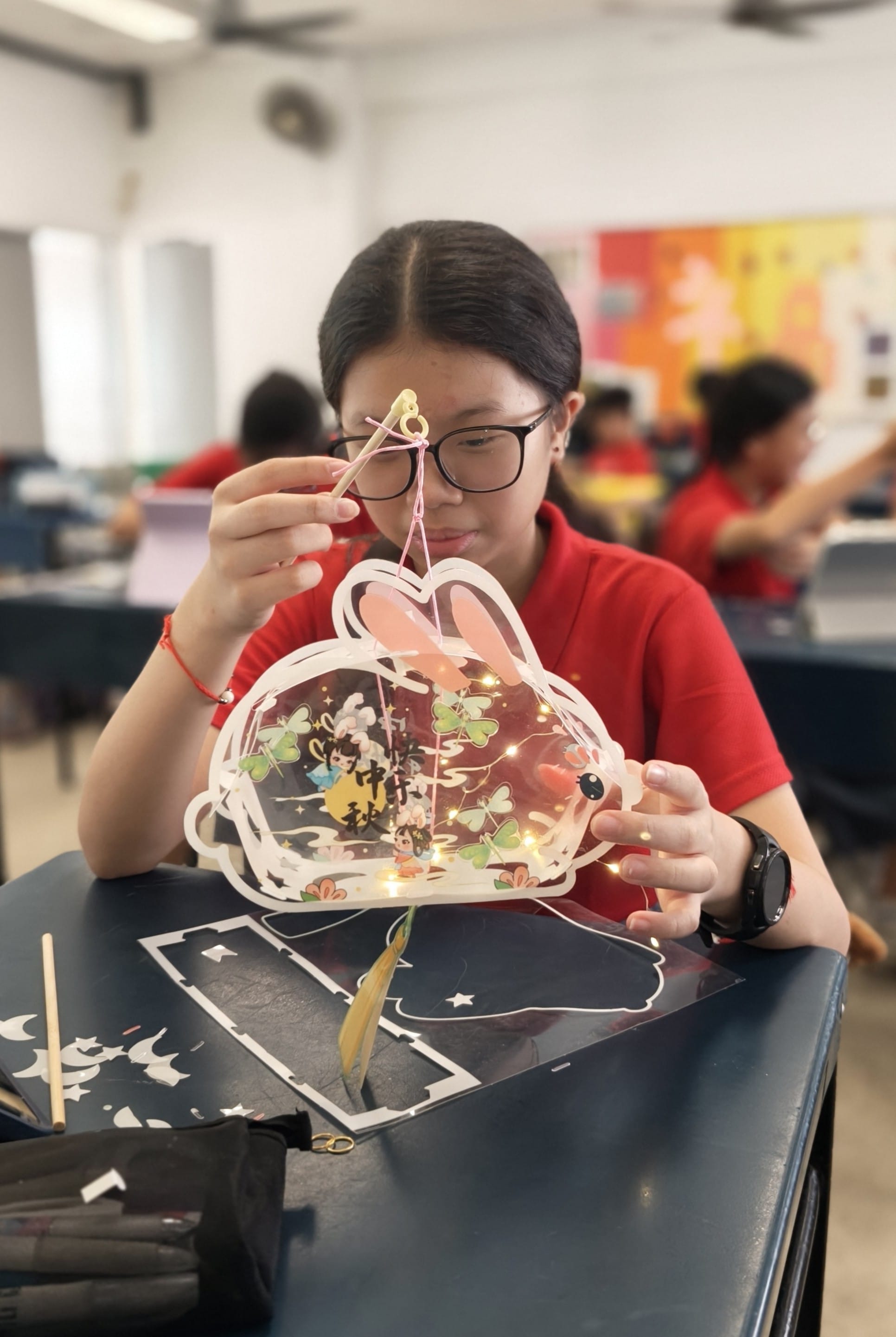 A female student wearing glasses and a red school uniform sits at a classroom desk, carefully threading string through a rabbit-shaped transparent lantern lit with fairy lights inside. The lantern features a Mid-Autumn Festival design with butterflies and Chinese characters. Other students work in the background.